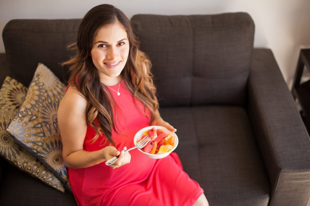 High angle view of a young pregnant woman sitting in the living room and eating some fruit from a bowl